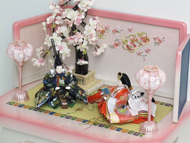 女雛はピンク、男雛は黒を主体とした松竹梅模様の西陣織り衣装を着せ付けたお雛さまをかわいいホワイトピンク収納台に桜で飾りました。