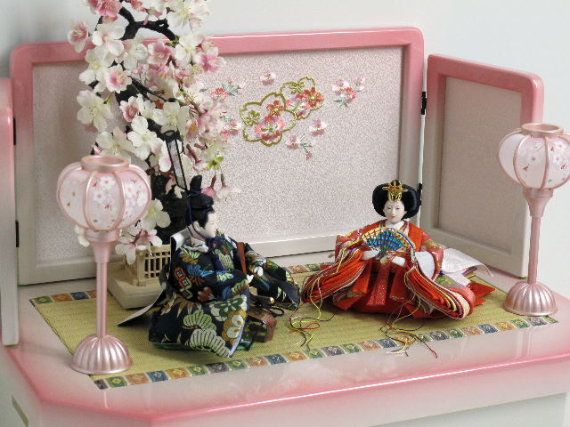 女雛はピンク、男雛は黒を主体とした松竹梅模様の西陣織り衣装を着せ付けたお雛さまをかわいいホワイトピンク収納台に桜で飾りました。