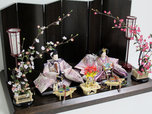淡い紫地に紫の桜を織り込んだ優雅で高貴なお雛さまを木目茶塗り台・六曲屏風と両脇からの紅白梅で引き立たたせました。